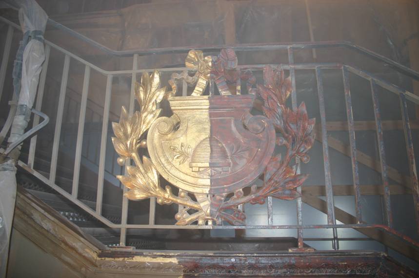 Zabytkowy, zdobiony metalowy herb na balustradzie schodów, który został częściowo wyczyszczony przy użyciu precyzyjnego czyszczenia suchym lodem. Lewa strona herbu jest jasna, metaliczna i czysta, pokazując jasny kolor mosiądzu lub brązu. Prawa strona herbu pozostaje ciemna, zabrudzona i zardzewiała